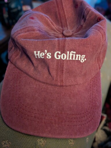 Distressed red baseball cap with white text that reads "He's Golfing."